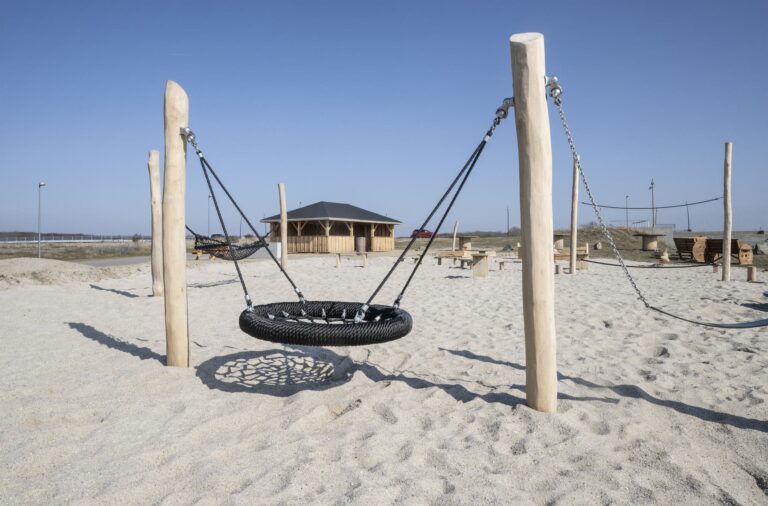 Fugleredegynge mellem stolper i træ i sandet ved Esbjerg Strand, med langhuset i baggrunden