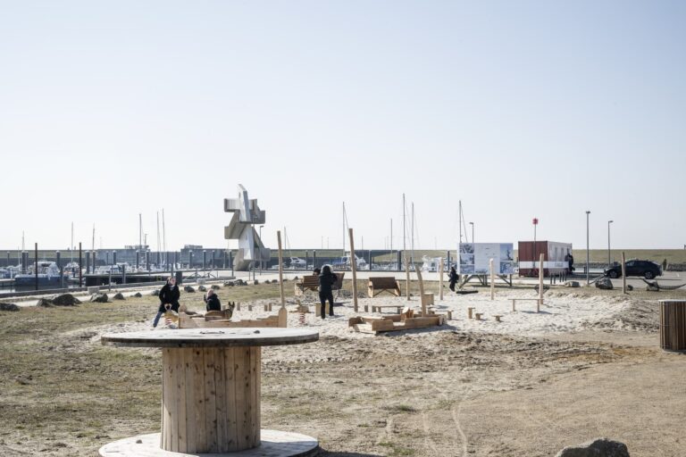 Kabeltromle i forgrunden og sandlegeområdet ved Esbjerg Strand, med havnen og skulpturen Mennesket ved Havet i baggrunden